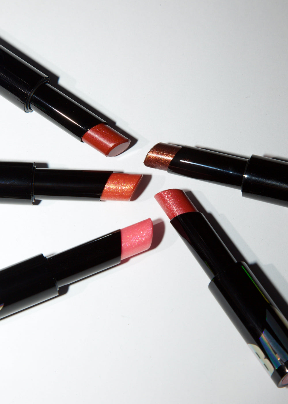 Four open lipsticks with black tubes arranged in a circle on a white background, showing different shimmering lipstick shades including orange, copper, pink, and red.