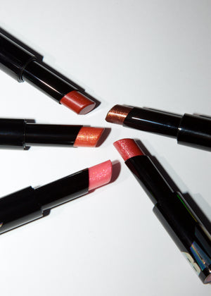 Four open lipsticks with black tubes arranged in a circle on a white background, showing different shimmering lipstick shades including orange, copper, pink, and red.